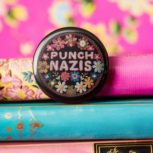 Round pinback metal button featuring an embroidered-style design on a black background. The center showcases bold, stitched-textured lettering in red, blue, and white that reads "PUNCH NAZIS." Surrounding the text is an intricate, folk-inspired floral pattern with multicolored flowers in shades of pink, orange, blue, and white, arranged symmetrically. A decorative zigzag border frames the edge of the button. The backside is silver metal with a secure pin closure.
